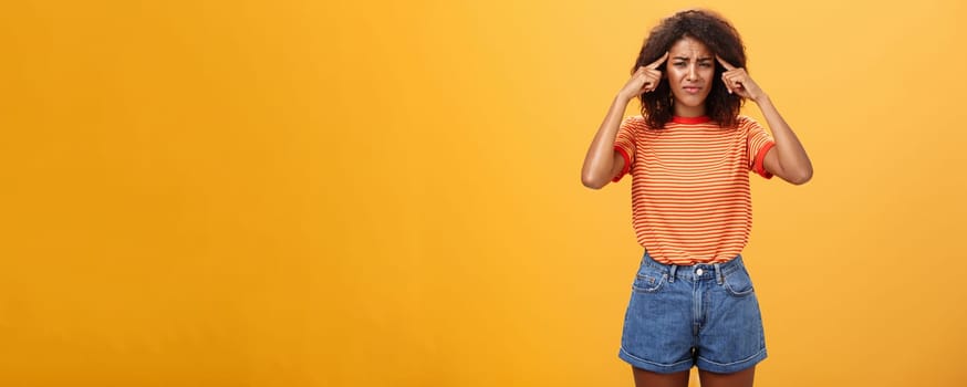 Girl cannot concentrate and think straight being loaded with bad thoughts looking concerned and intense, frowning holding fingers on temples suffering disorientation and migraine over orange wall. Emotions and health concept