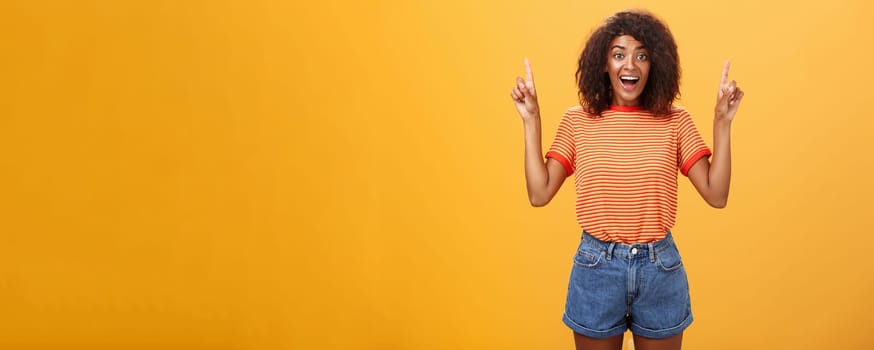 Charmed and impressed african american stylish woman seeing incredible and awesome item on sale raising hands and pointing up with broad grin being excited of new purchase over orange wall. Emotions and advertisement concept