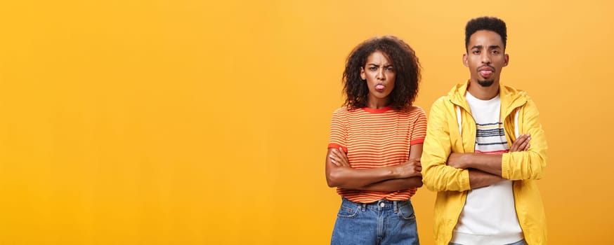 Indoor shot of african american siblings being displeased and annoyed showing bad tempber behaving childish sticking out tongue standing together with crossed hands on chest over orange background. Lifestyle.