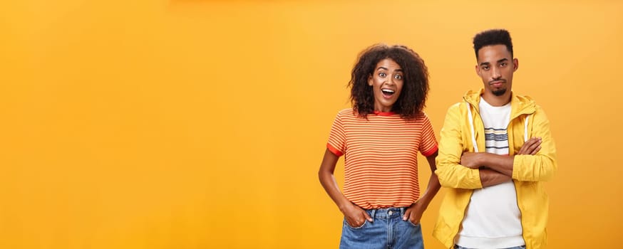 Two african american friends standing over orange background, girl thinks idea is awesome feeling excited and joyful while boyfriend being displeased and indifferent to unimpressive plan. Copy space