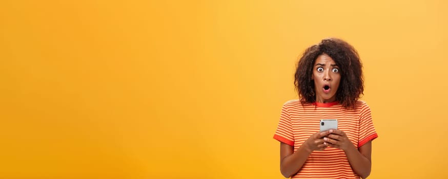 Portrait of shocked and concerned stunned dark-skinned woman with afro hairstyle popping eyes from concern and panic opening mouth holding smartphone revealing terrible information.