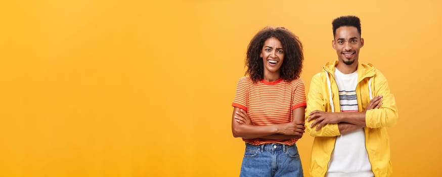 Portrait of surprised sarcastic african american woman with afro hairstyle standing with cute boyfriend crossing arms on chest laughing from scorn and fun over orange background. Copy space