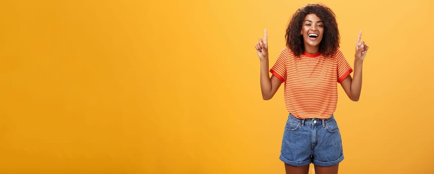 Woman feeling amused and entertained. Portrait of happy carefree stylish African-American girl with afro hairstyle laughing out loud joyfully pointing up with raised arms over orange wall. Emotions concept