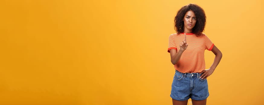 You better not. Dissatisfied bossy serious-looking african american curly-haired female head manager in t-shirt frowning shaking index finger in forbid or stop gesture scolding dog over orange wall. Body language concept