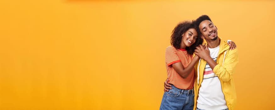 They love each other. Portrait of two charming african american man and woman in relationship hugging with heartwarming smile touching hands smiling gently posing against orange background.