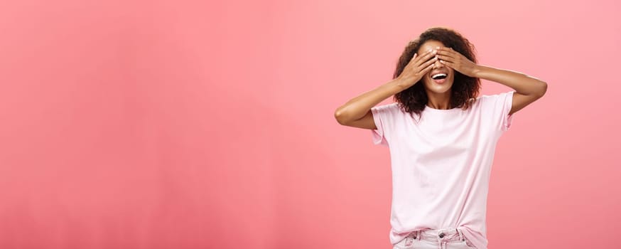 Portrait of charming joyful dark-skinned playful woman with curly hair in t-shirt closing eyes and counting ten with broad happy smile playing hide n seek or waiting for surprise over pink background. Lifestyle.