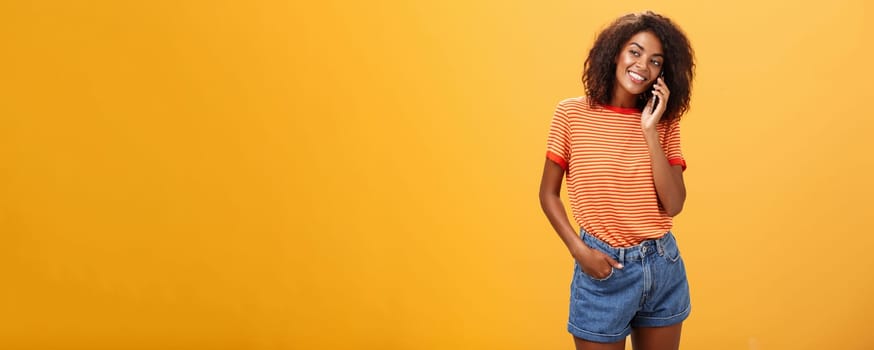 Stylish dark-skinned girl making casual phone call to friend telling all details of after romantic date standing pleased and carefree over orange background in striped t-shirt gazing left with grin. Lifestyle.