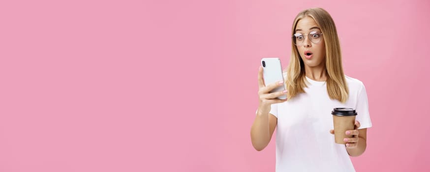 Woman drinking coffee being shocked by received message reacting on stunning news folding lips gasping looking astonished and impressed at smartphone screen holding paper cup, posing over pink wall. Technology concept