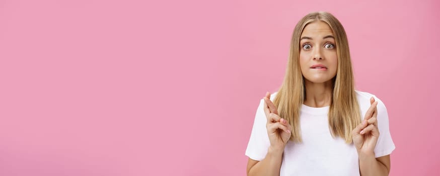 Excited and thrilled attractive emotive fair-haired female with pimples on forehead biting lower lips crossing fingers for good luck making wish praying desire become reality over pink wall. Emotions and body language concept