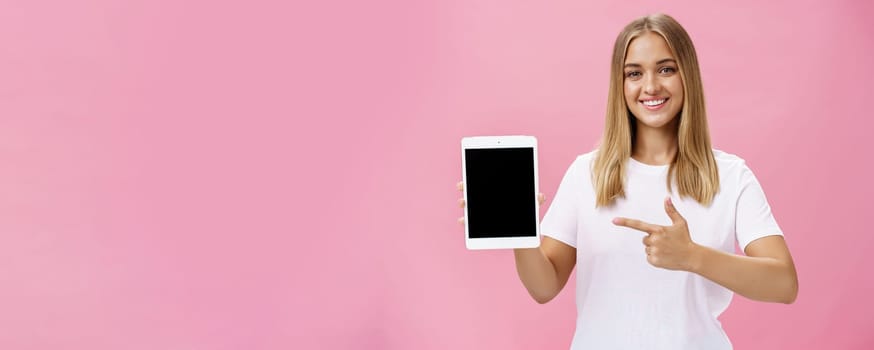 Girl recommends use only this digital tablet. Charismatic young female with fair hair tanned skin and no makeup in white t-shirt pointing at gadget screen and smiling friendly at camera over pink wall. Technology and advertisement concept