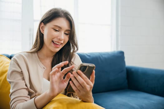 In a relaxed home setting a woman on a sofa happily talks on her phone typing messages. Engaged in successful communication and technology she embodies relaxation. The woman is relaxing.