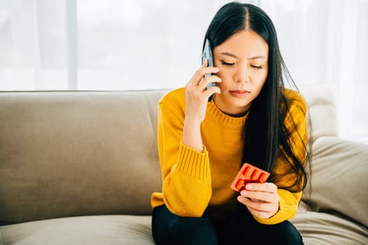 Highlighting consultation, Asian woman takes medicine consults doctor via phone while on couch at home. Depicting healthcare quarantine and social distancing amid coronavirus.