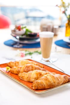 A delightful breakfast with croissants and coffee on a marble table. Croissants are golden brown on an orange plate, coffee with froth.