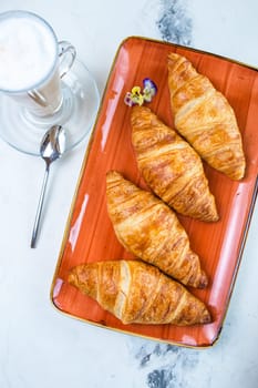 A delightful breakfast with croissants and coffee on a marble table top view. Croissants are golden brown on an orange plate, coffee with froth.