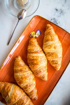 A delightful breakfast with croissants and coffee on a marble table top view. Croissants are golden brown on an orange plate, coffee with froth.