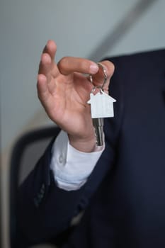 The image shows a persons hand holding a silver house key with a house-shaped keychain. The key is isolated on a white background.