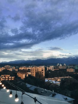 A stunning cityscape with mountains in the backdrop, surrounded by green trees under a blue sky with white clouds.