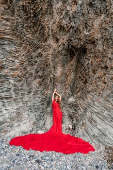 woman sea red dress. Woman with long hair on a sunny seashore in a red flowing dress, back view, silk fabric waving in the wind. Against the backdrop of the blue sky and mountains on the seashore