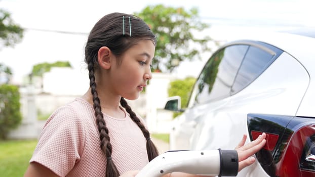 Happy little young girl learn about eco-friendly and energy sustainability as she recharge electric vehicle from home EV charging station. EV car and sustainable future generation concept. Synchronos