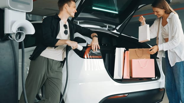 Young couple with shopping bag travel by EV electric car to shopping center parking lot charging in green city urban sustainability lifestyle by clean rechargeable energy of electric vehicle innards
