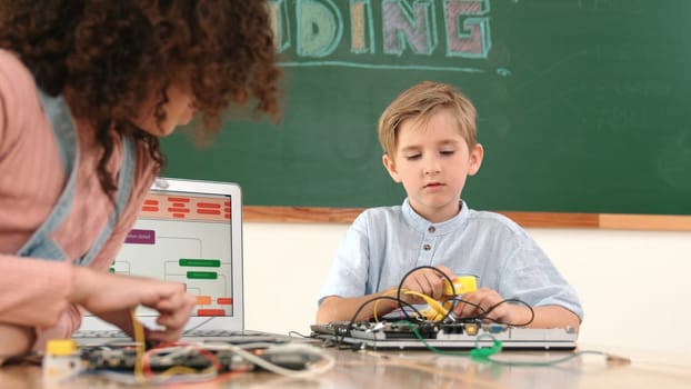 Caucasian smart boy fixing main board by using screwdriver at stem technology lesson while smiling student working together to inspect electronic machine at table with laptop display code. Pedagogy.
