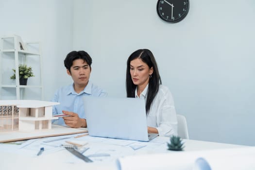 Professional male asian architect using ruler to measure house model length while young beautiful caucasian colleague using laptop to analyzed data on meeting table with house model. Immaculate.