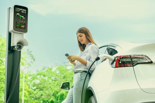 Young woman using smartphone online banking application to pay for electric car battery charging from EV charging station during vacation holiday road trip at national park or summer forest. Exalt