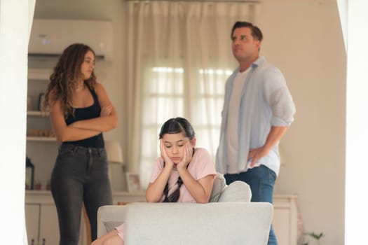 Annoyed and unhappy young girl sitting on sofa trapped in middle of tension by her parent argument in living room. Unhealthy domestic lifestyle and traumatic childhood develop to depression.Synchronos