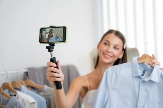 Young woman making fashion video content for social media. Blogger smiles to camera using selfie stick and light ring while making persuasive online clothing sell to audience or followers. Unveiling