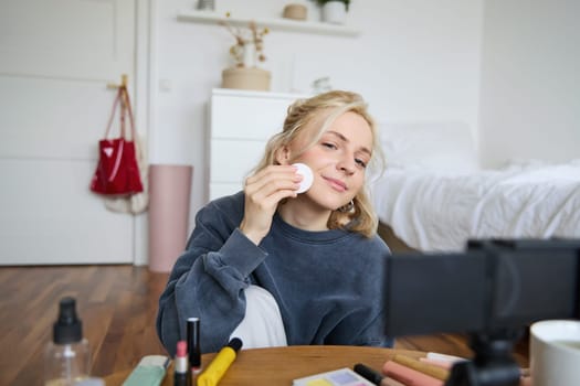 Portrait of young woman promoting beauty product, applies makeup in front of camera, recording video for her vlog, creating content for social media, sitting in a room on floor.