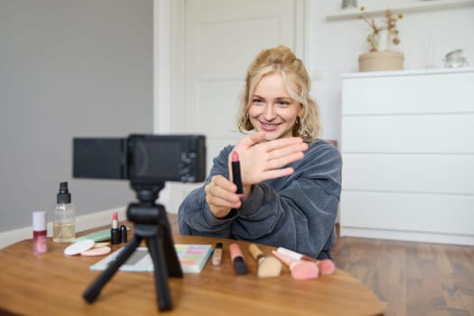 Portrait of young blond woman, teenage girl records video for her social media account, shows makeup on camera, recommends lipstick to online followers, creates content in her room.