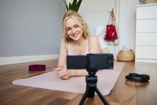 Portrait of young woman, sports vlogger, fitness instructor recording video of herself showing workout exercises, using digital camera, lying on yoga rubber mat.