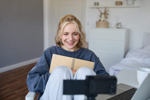 Portrait of blond smiling woman, records video on digital camera how she writes in notebook, talks to followers, doing lifestyle blog content in her room.