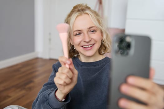 Image of stylish young woman, social media influencer, taking pictures on mobile phone, doing makeup tutorial for followers online, recording video vlog in her bedroom, showing brush.