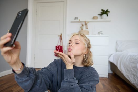 Image of young woman sits on floor in her bedroom, takes selfies on smartphone, sends air kisses at mobile phone camera, video chats online using app.