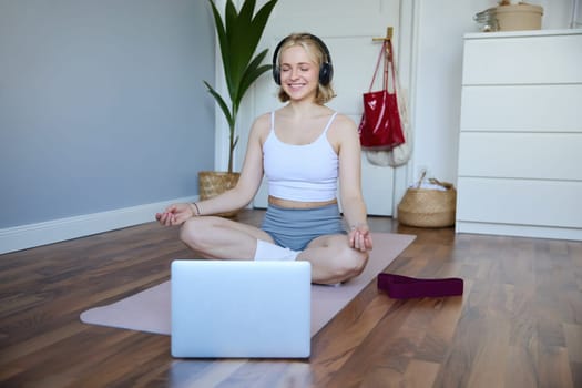 Portrait of young and relaxed woman using laptop and wireless headphones while meditating, practice yoga at home on rubber mat, sitting in asana.