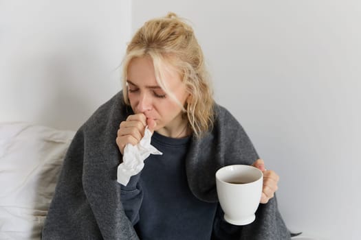 Concept of flu and quarantine. Close up portrait of young woman feeling sick, caught a cold, sneezing and blowing nose in napkin, holding warm tea in hand.