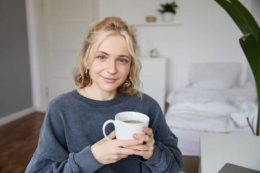 Portrait of smiling beautiful young woman, sitting in bedroom with cup of tea, resting at home alone, enjoys her weekend indoors.