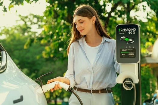 Young woman recharging battery for electric car during road trip travel EV car in natural forest or national park with sunrise lighting. Eco friendly travel during vacation and holiday. Exalt