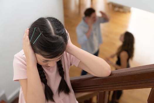 Stressed and unhappy young girl cover her ears blocking sound tension by her parent argument from the stair. Unhealthy family and domestic violence lead to traumatic childhood and anxiety. Synchronos