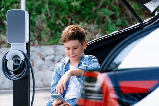 Little boy sitting on car trunk while recharging eco-friendly electric car from EV charging station. EV car road trip travel concept for alternative transportation powered sustainable energy.Perpetual