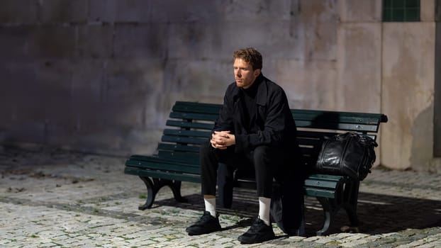 A man sits on a bench in the dark. He is wearing a black coat and has a backpack on his lap. The scene is quiet and peaceful, with the man looking off into the distance