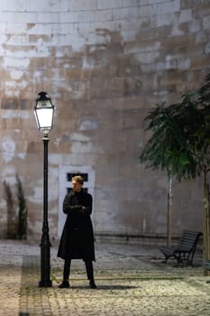 A man stands in front of a street lamp, looking at the camera. The scene is set in a city at night, with a bench nearby. The woman is wearing a black coat and she is waiting for someone or something