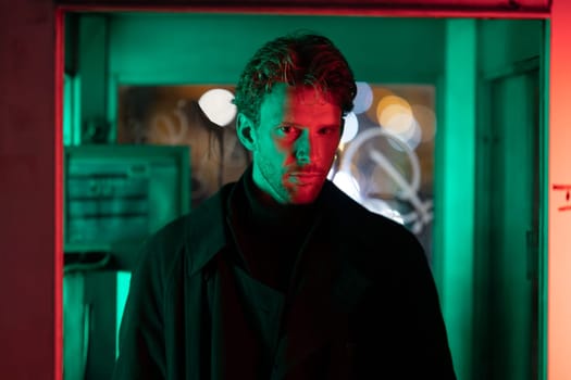 A man is standing in front of a neon sign. The sign is green and red. The man is wearing a black coat and a black hat