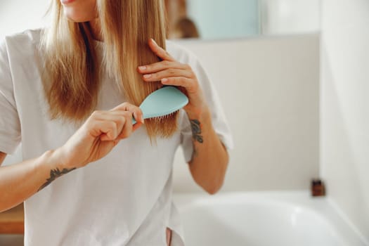 Woman standing in front of mirror at bathroom and combing her hair with brush after shower