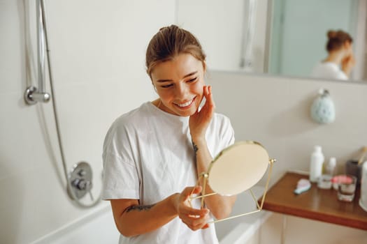 Smiling young woman looking in small round mirror and checking skin while standing in bathroom