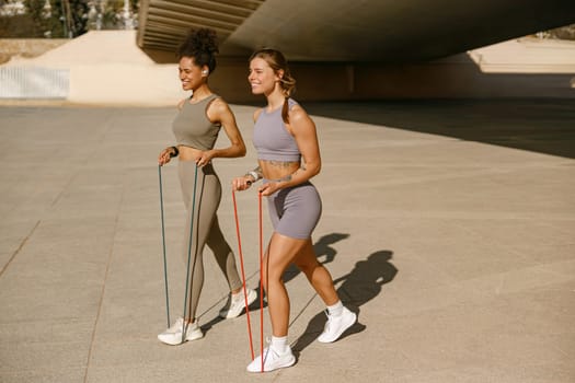 Two young smiling women makes fitness workouts with resistance band on morning city background