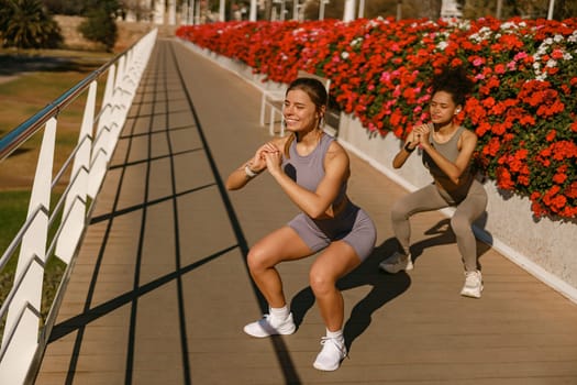 Two smiling fit women doing workout and squatting together outdoors in the city