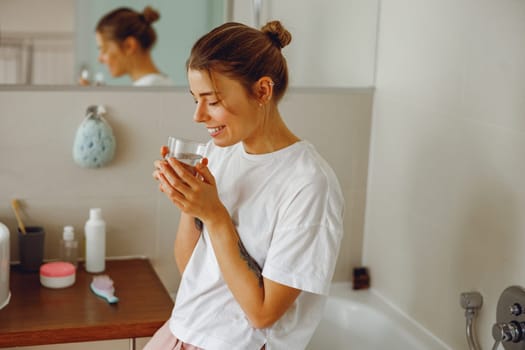 Young beautiful woman drinking glass of water in the bathroom at home. Beauty treatment