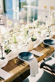 Black plates stand on a wooden table near white bouquets of flowers and candles on a narrow tablecloth. High quality photo